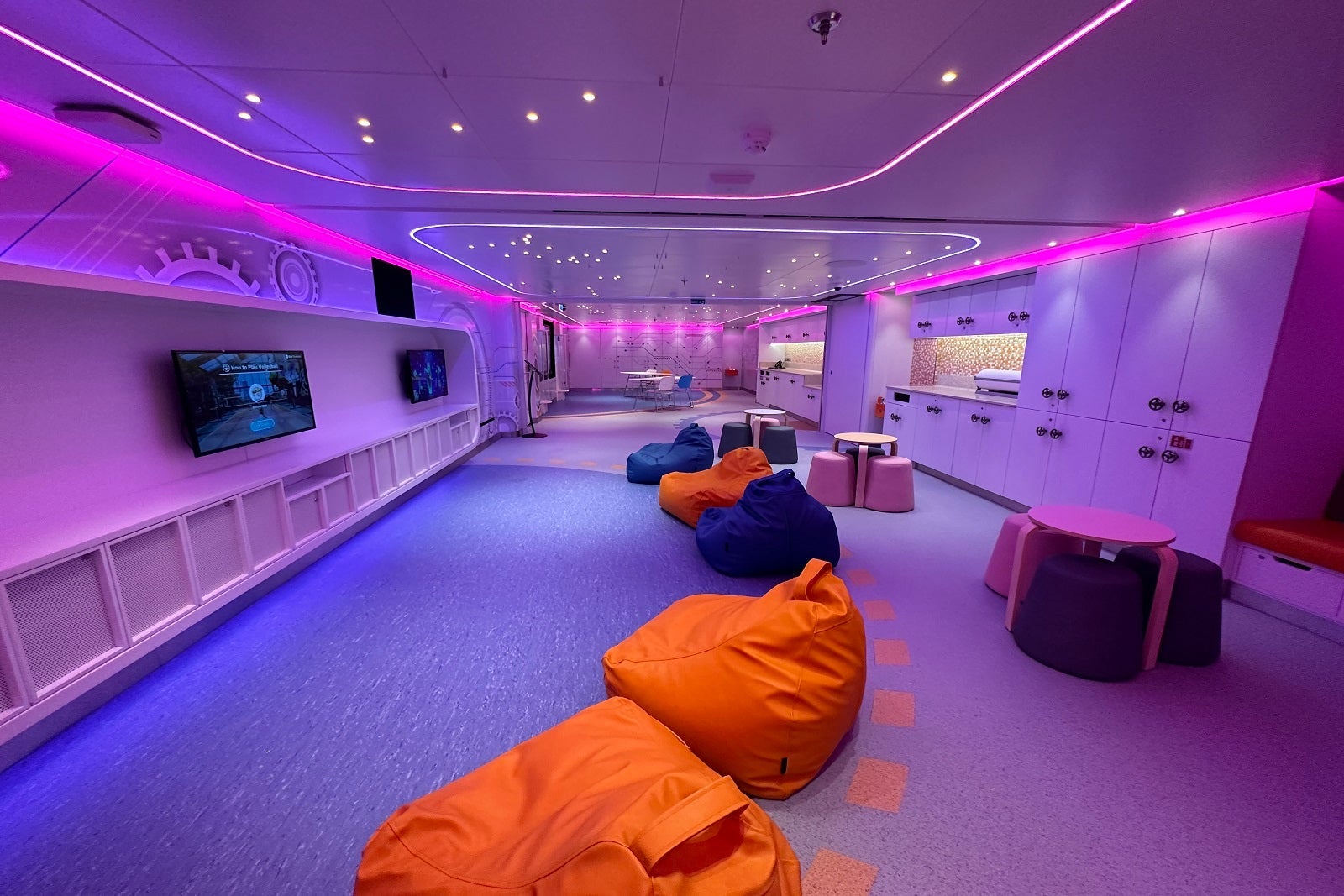 A kids club space on a cruise ship with purple lighting, orange and blue beanbag chairs and wall-mounted TVs