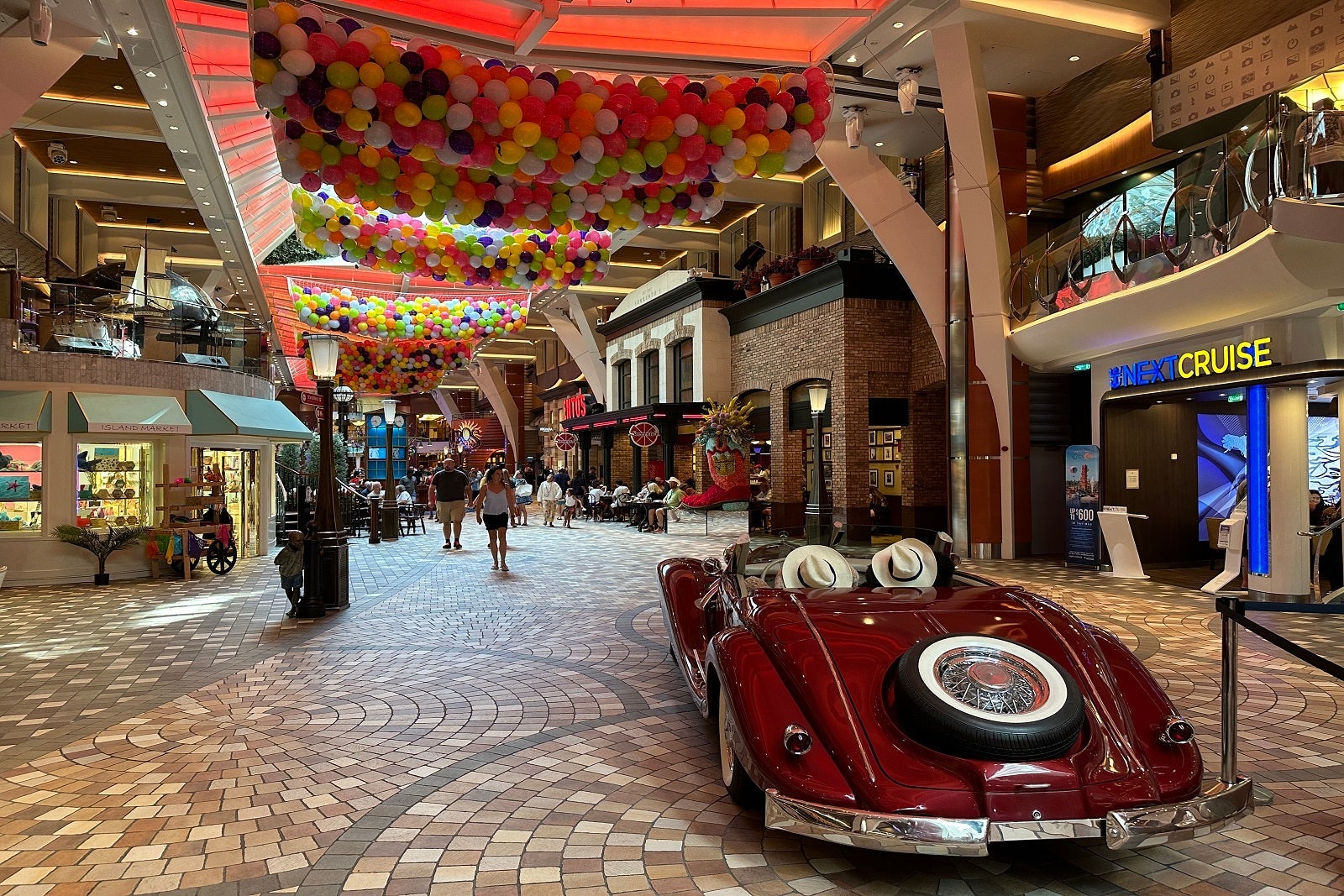 A classic car parked along an indoor cruise ship promenade with a balloon drop setup overhead