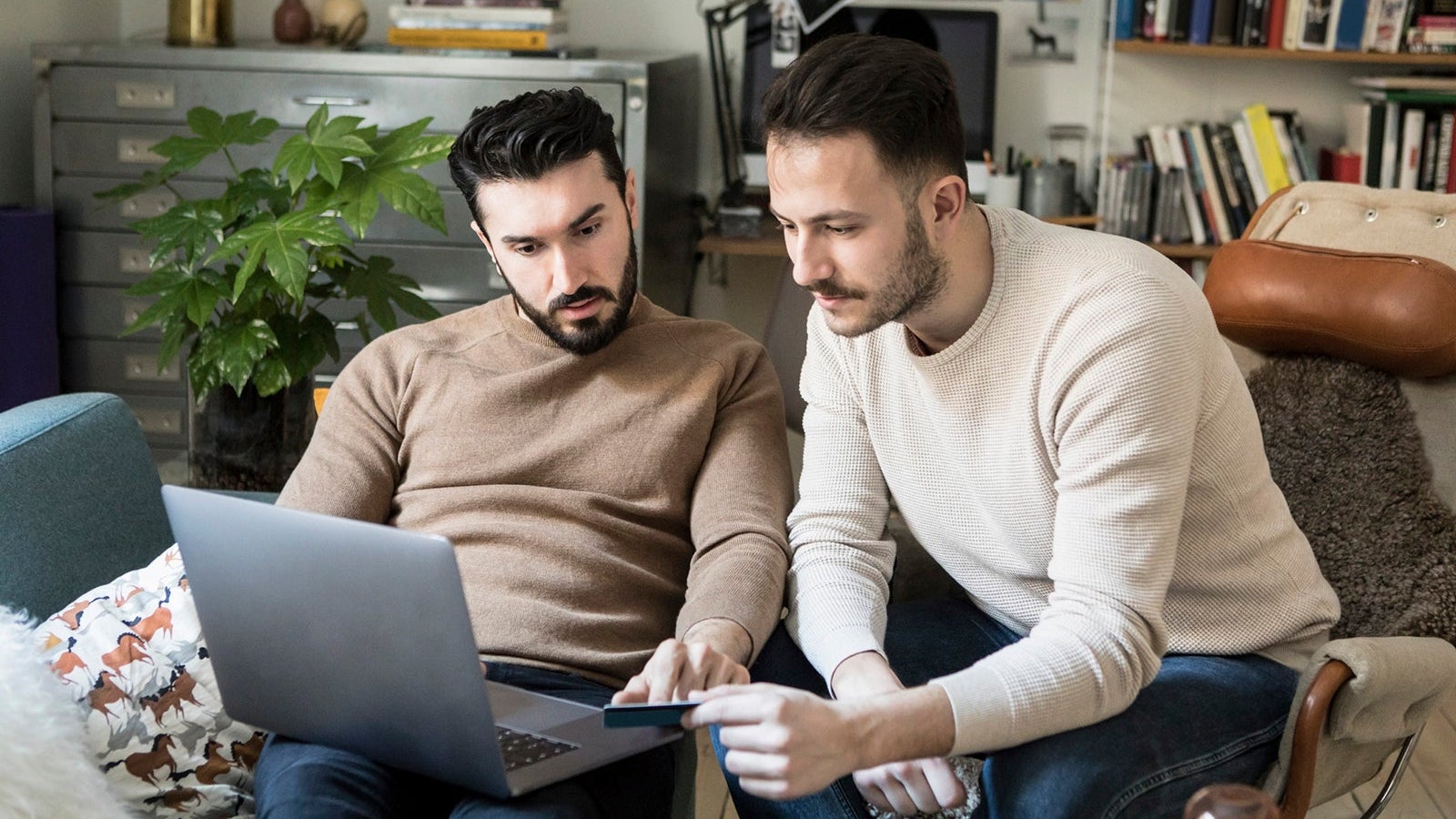 Gay couple shopping online with credit card through laptop while relaxing on chairs at home