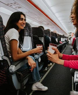 Two woman drink coffee and have a light hearted chat on a plane