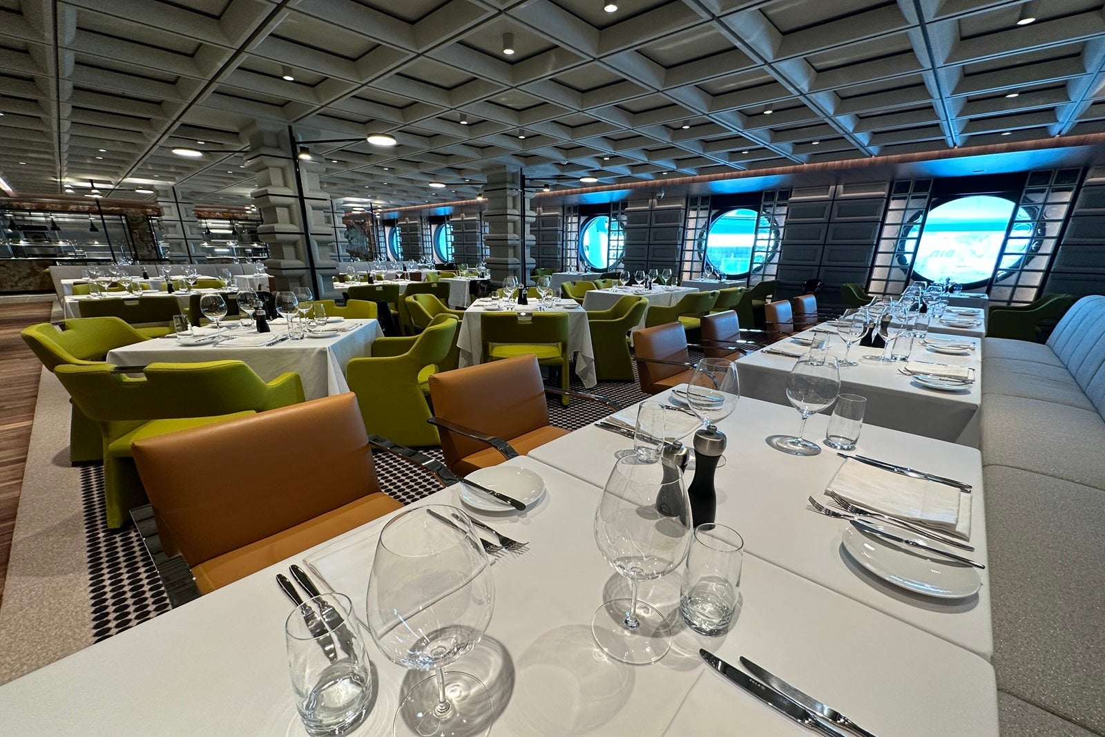A midcentury modern restaurant setup with a wooden floor, green and brown chairs and white tablecloths with place settings on a cruise ship