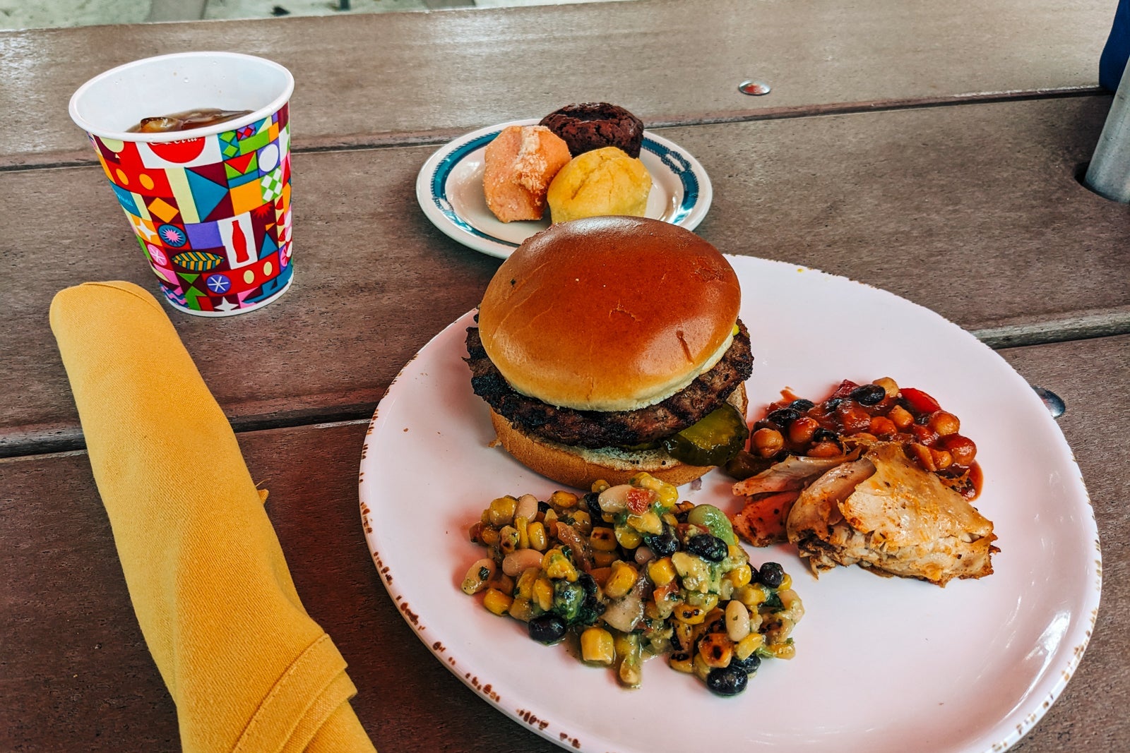 Beach lunch on Castaway Cay -- white plate with burger, bean salad and chicken, plus an extra plate of dessert and a drink in colorful cup