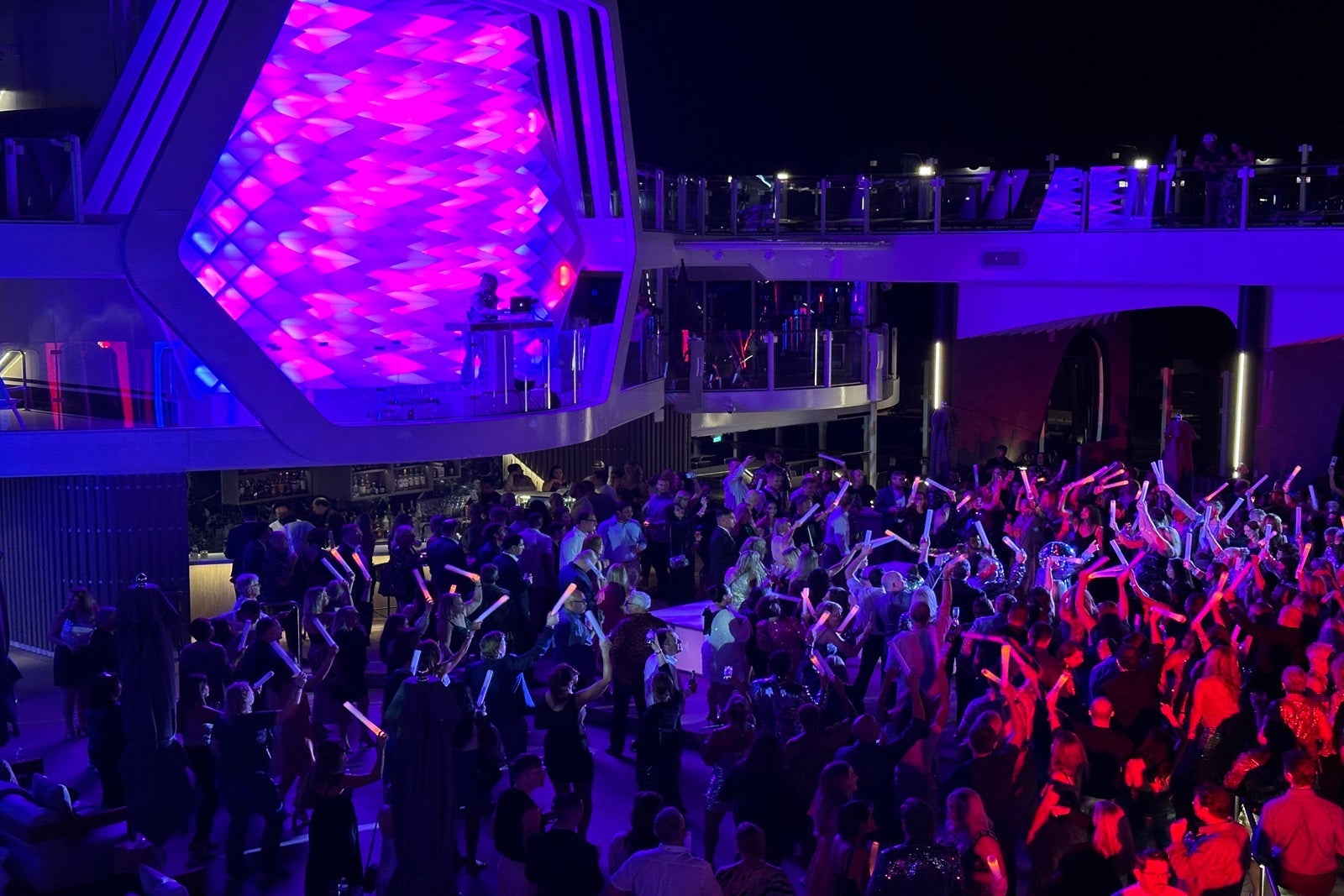 A dancing crowd gathered in front of a DJ booth for a party on the pool deck of a cruise ship at night
