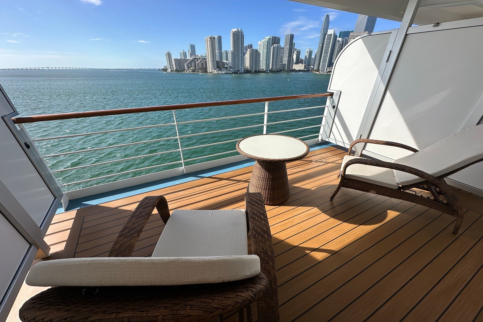 A cruise ship balcony with two padded chairs, a coffee table and a railing overlooking teal water and the Miami skyline