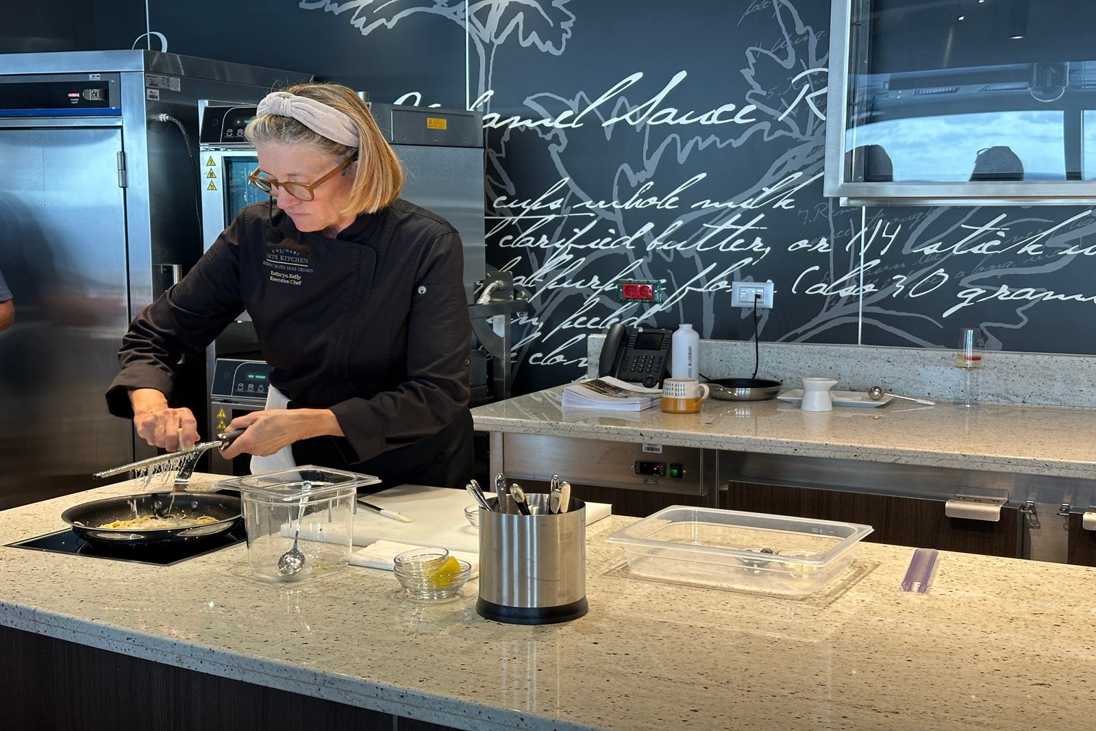 A chef stands at a table demonstrating how to prepare a dish during a cooking class on a cruise ship