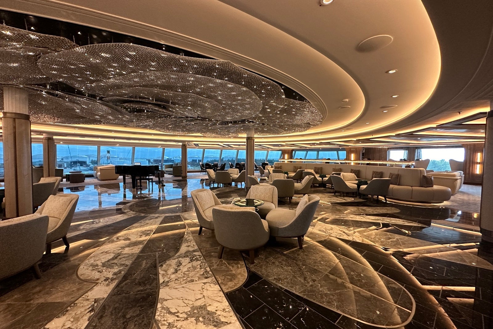 A large sitting area with a marble dance floor, gray and black carpeting, gray tables and chairs, a black piano, sparkling light fixtures and floor-to-ceiling windows