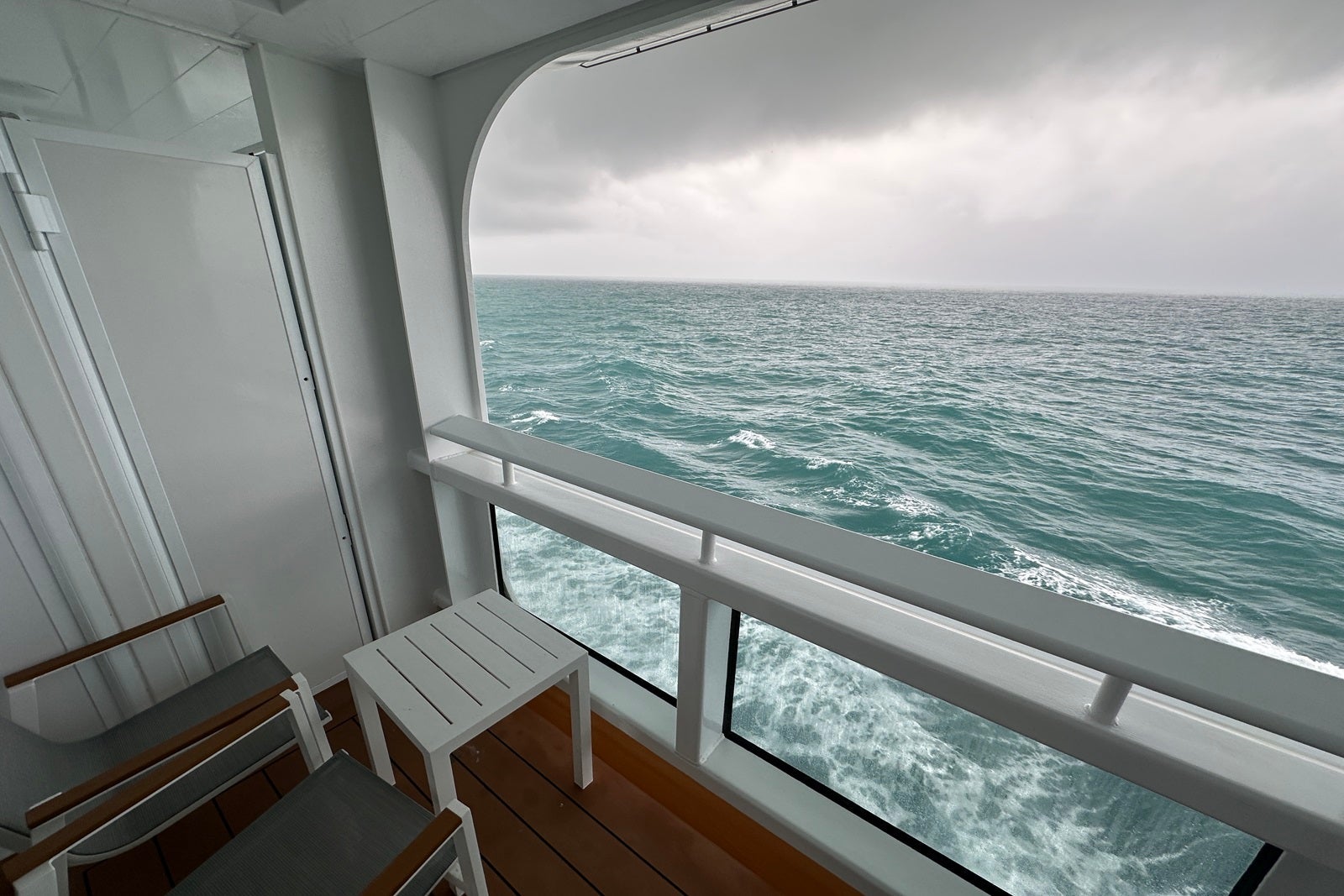 Angled shot of the view from a cruise ship balcony with teal waves, a railing and a small table with two chairs