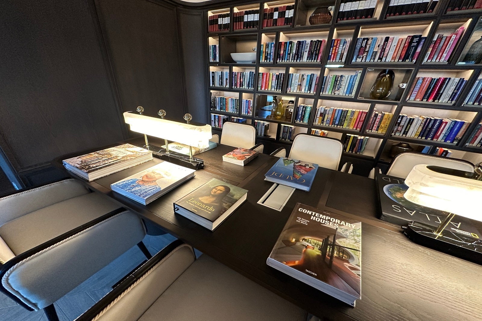 A dark library with a wall of books and a reading table with coffee table books on top of it, as well as reading lamps
