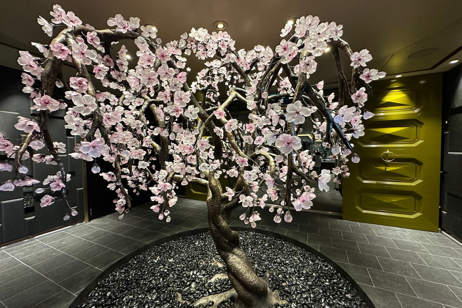 A Japanese cherry blossom tree sculpture adorned with pink glass flowers stands outside a cruise ship restaurant