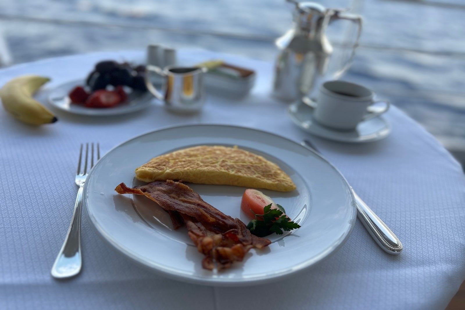 A table set with a white cloth, silverware, a coffee caraffe and a plate with an omelet, bacon and a strawberry