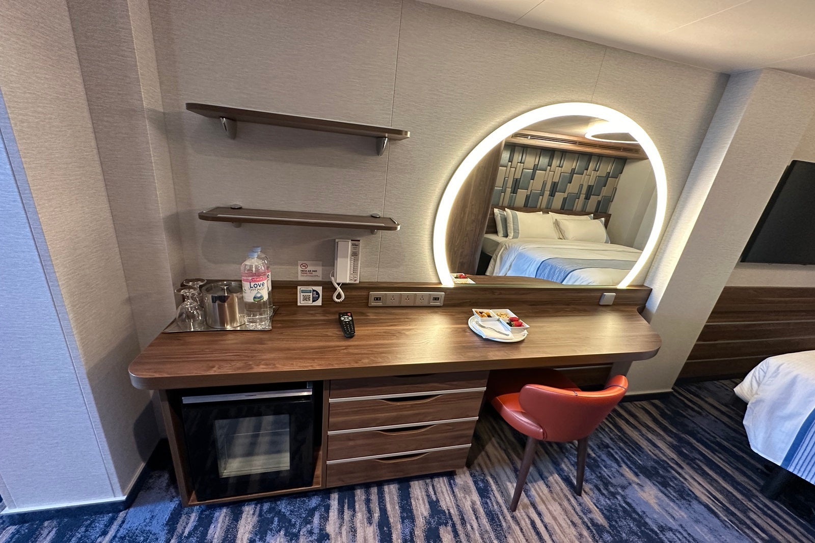 A wooden vanity with shelves, drawers, a mini-fridge, a chair bottles of water, electrical outlets and a rounded mirror with lighting