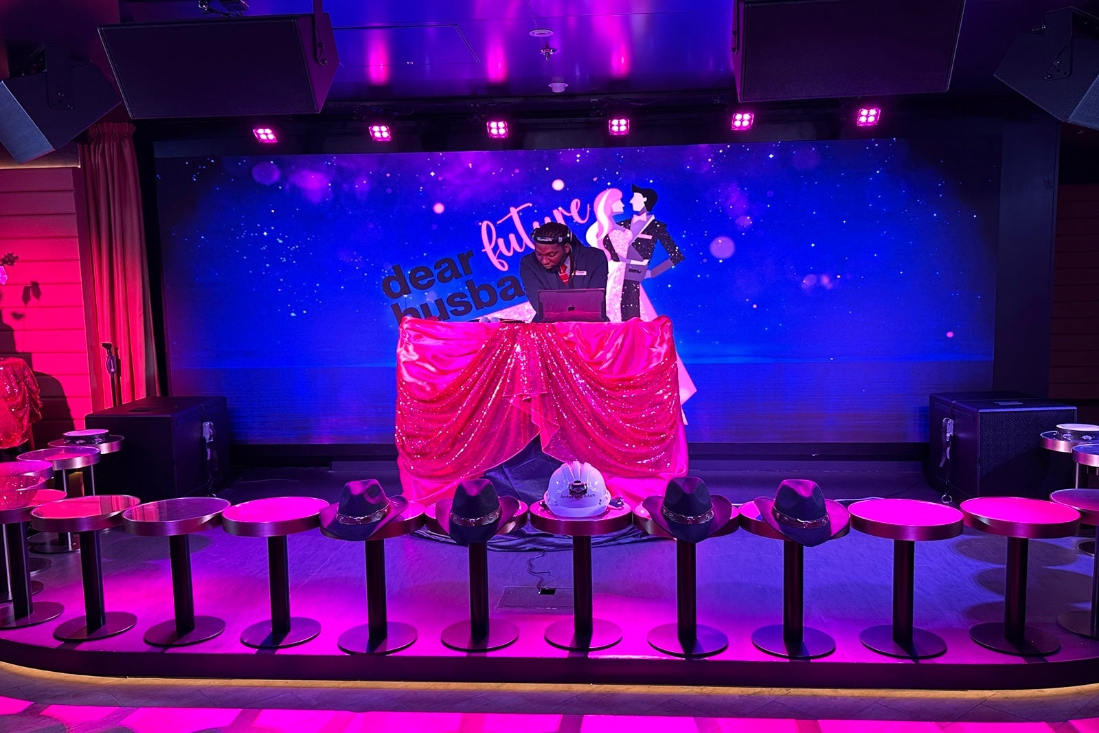 A DJ spins tracks at a table with a pink tablecloth and a blue LED screen in the background