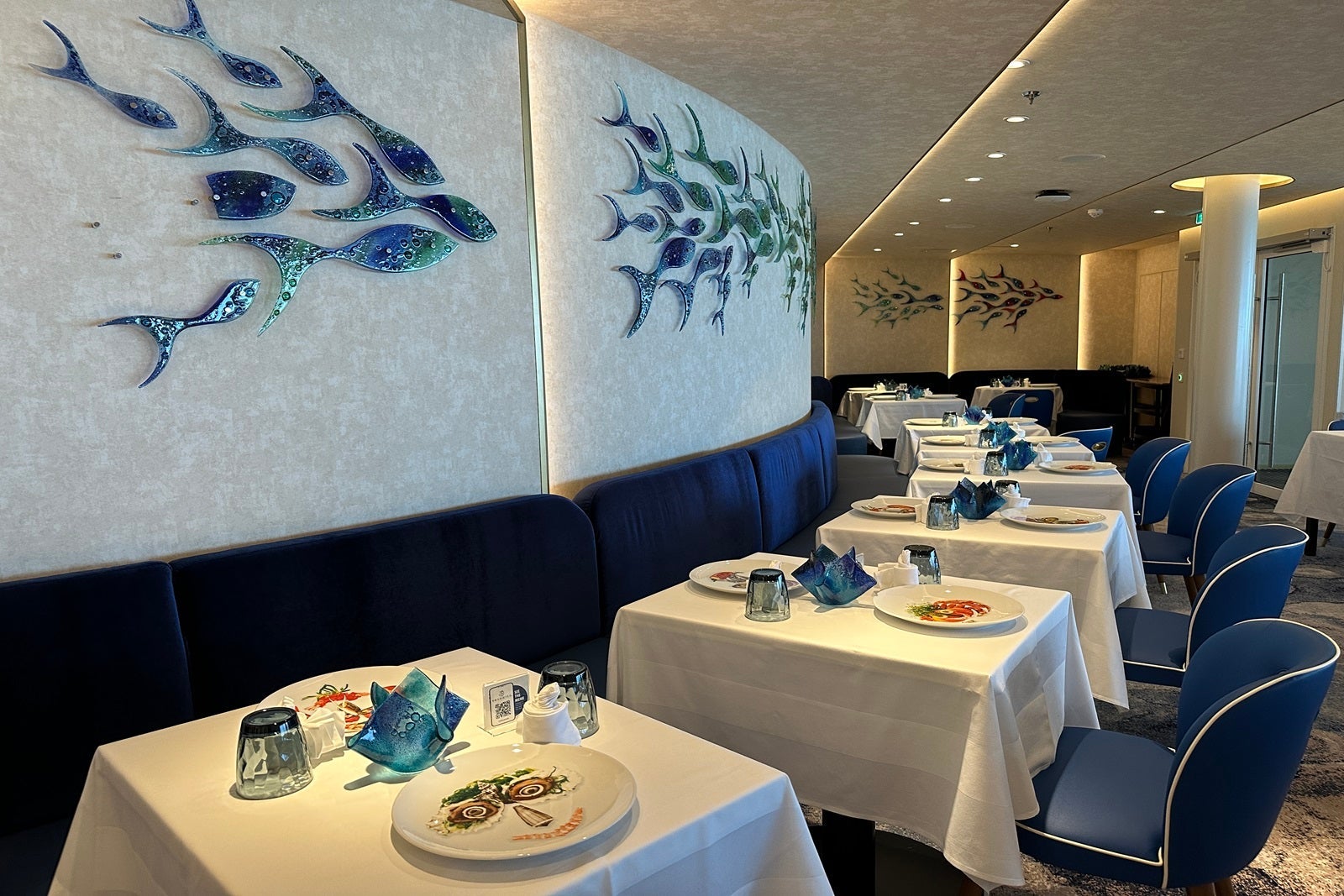 In front of a wall of glass ornamental fish, a row of tables with white tablecloths, set with dishes that have faces on them