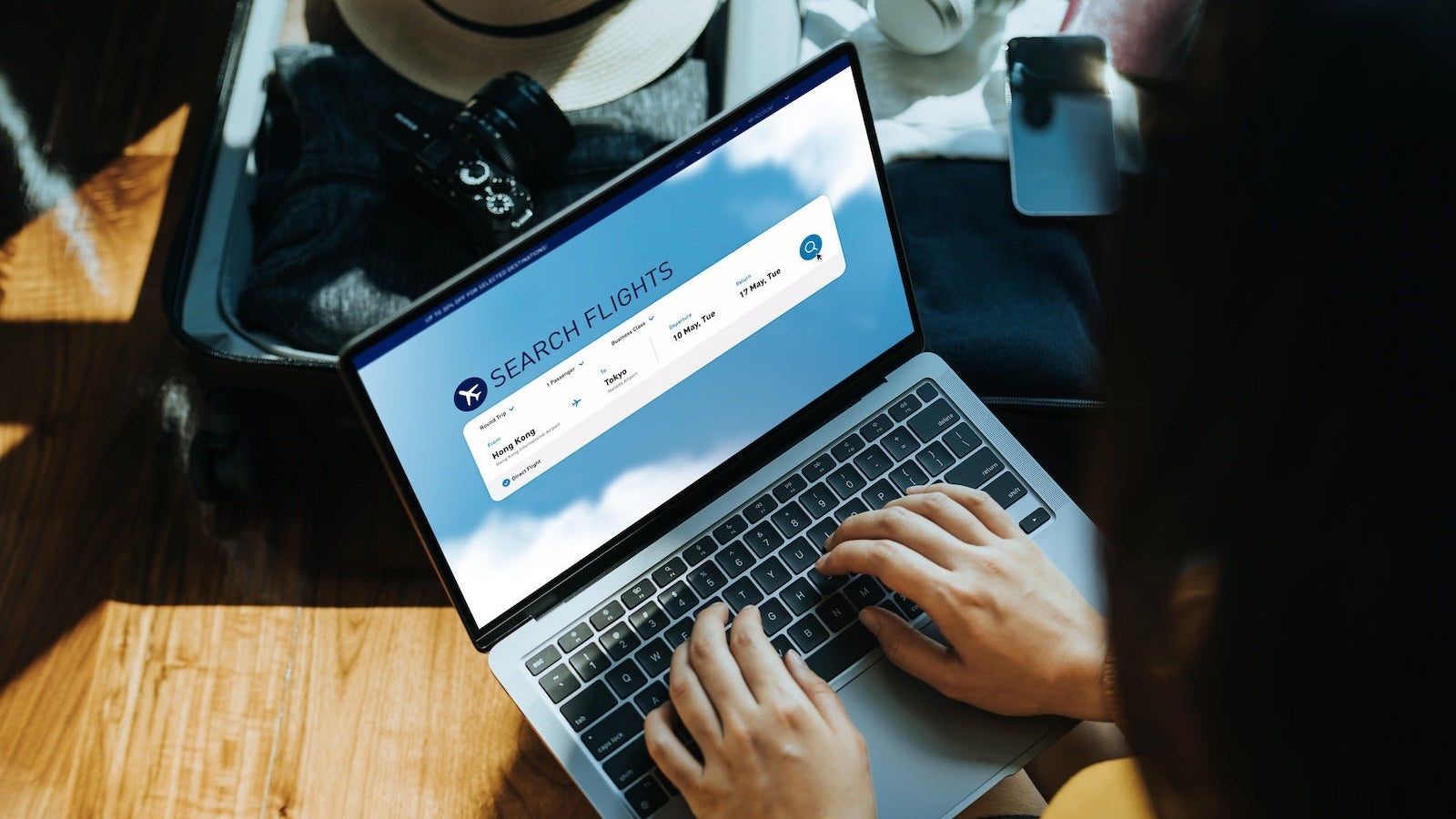Young Asian woman shopping online for flight ticket on airline website with laptop at home, sitting next to suitcase with sun hat, camera, clothings, headphones, smartphone and passport. Getting ready for vacation. Travel and vacation concept