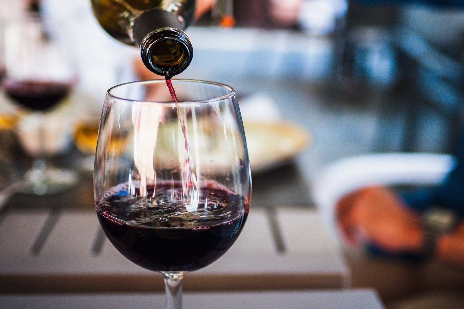 Red wine pouring into a clear glass from a bottle at the top of the frame with a blurred table and food in the background