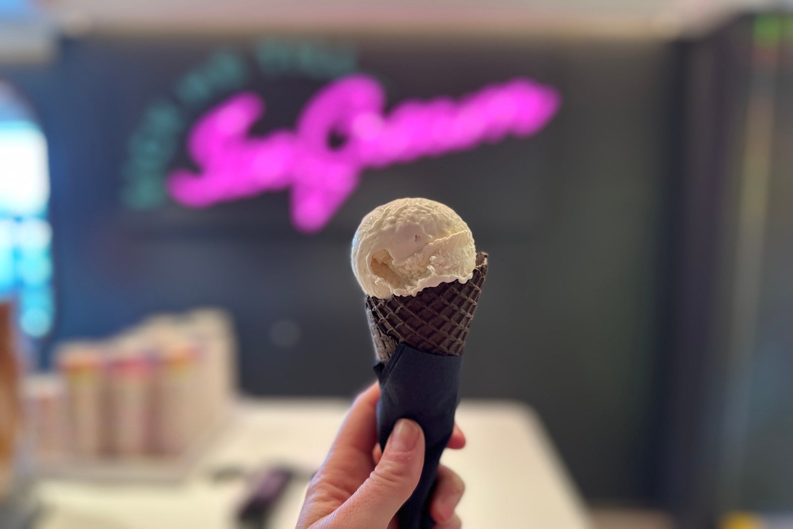 A hand holding an ice cream cone in front of a blurred neon sign