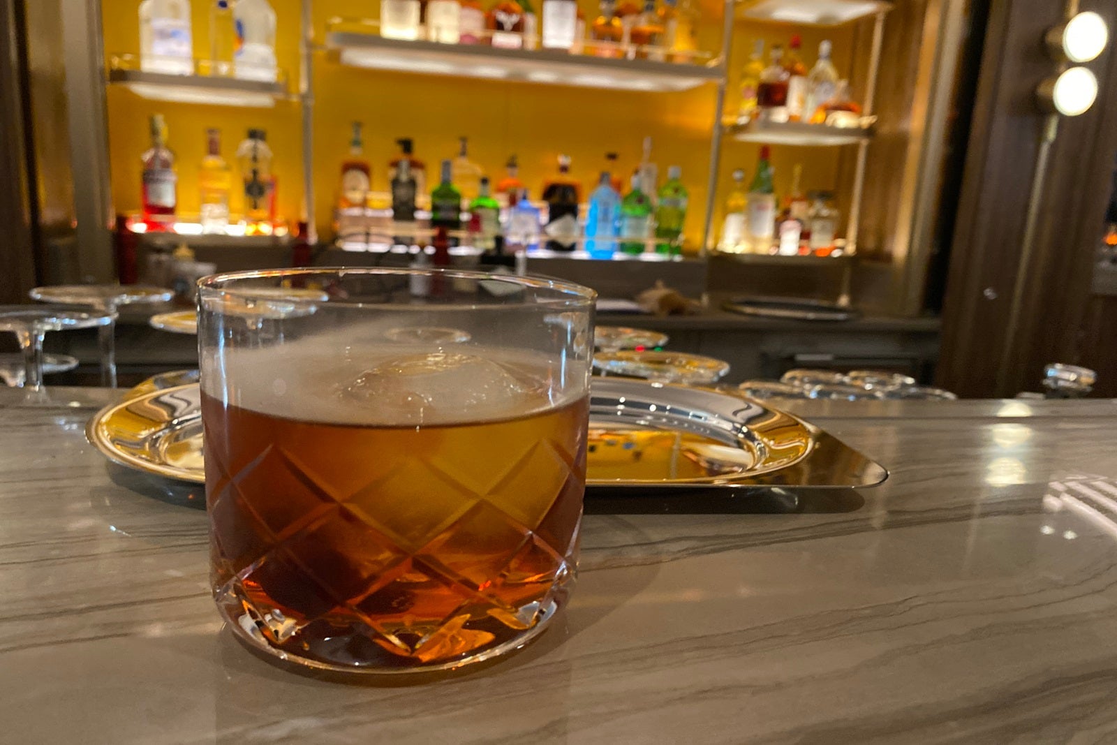 A cocktail sits in a glass on top of a bar with liquor bottles in the background