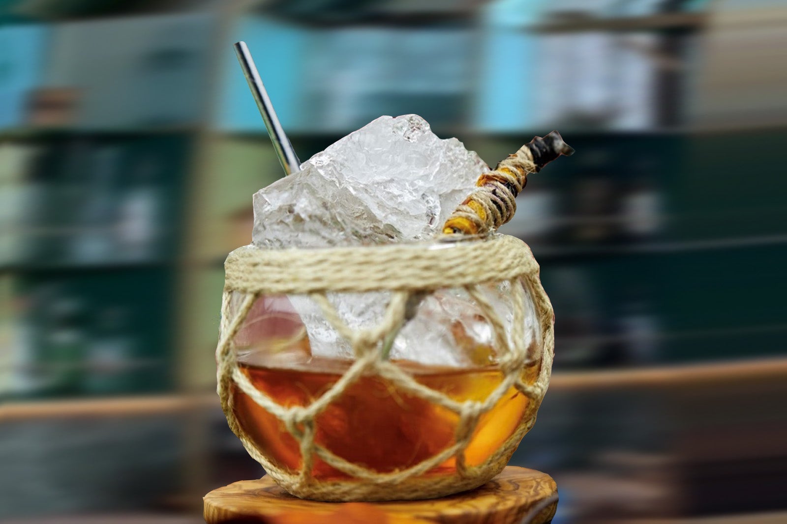 A cocktail in a spherical glass covered in twine with a metal straw, a large chunk of ice and a piece of burning sage