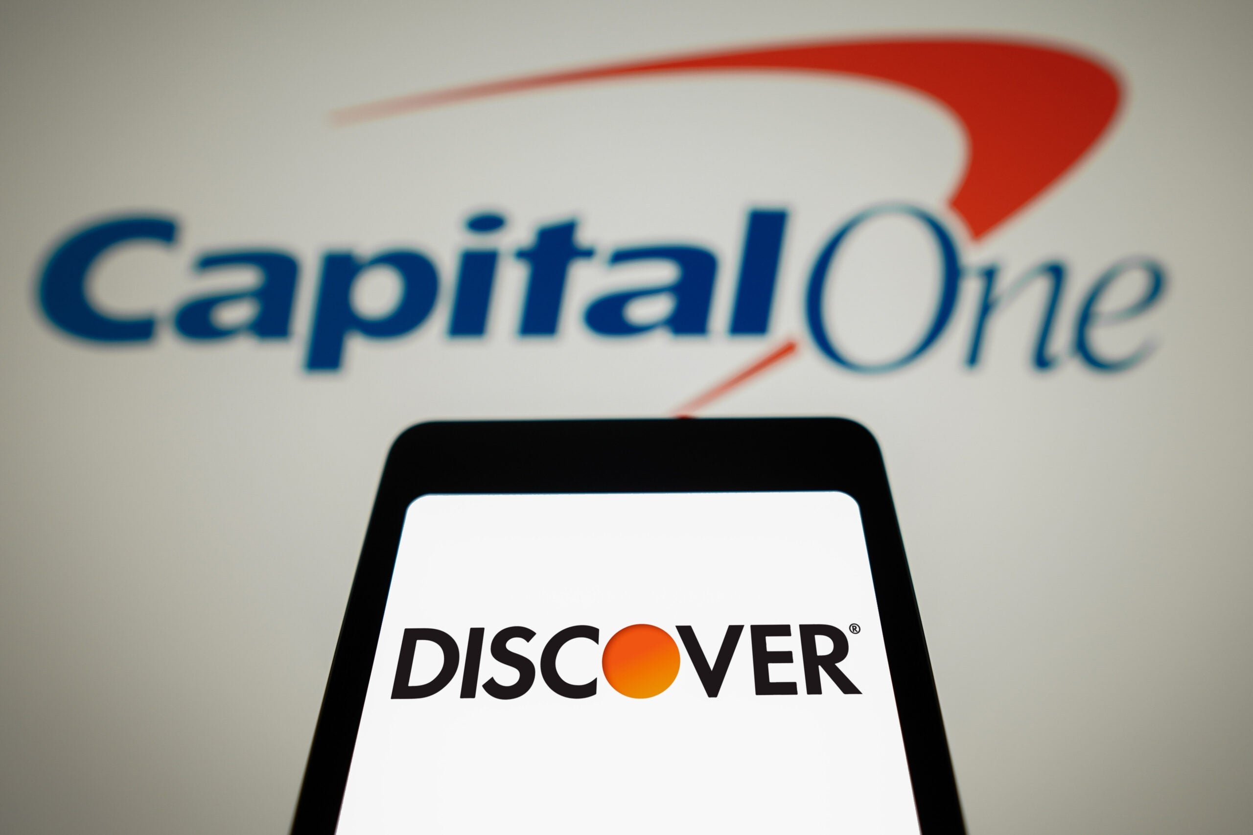 In this photo illustration, the Discover Financial Services logo is displayed on a smartphone screen with the logo Capital One Financial in the background.
