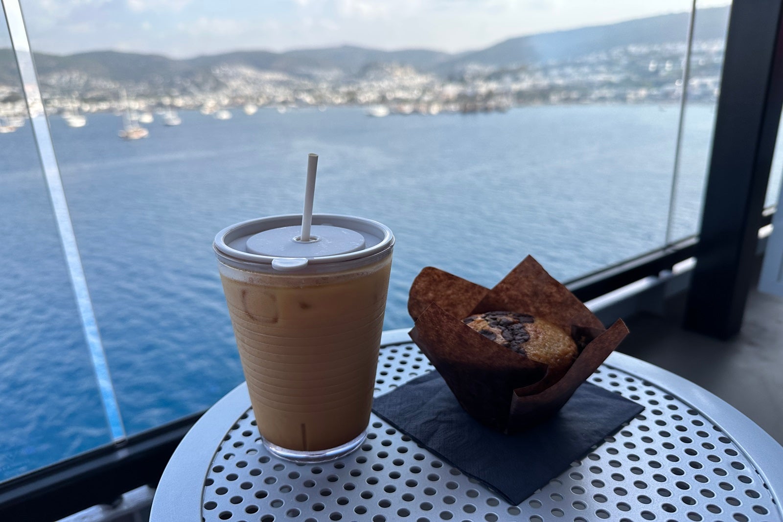 A cup of iced coffee and a muffin wrapped in brown paper sit on a metal table overlooking the ocean