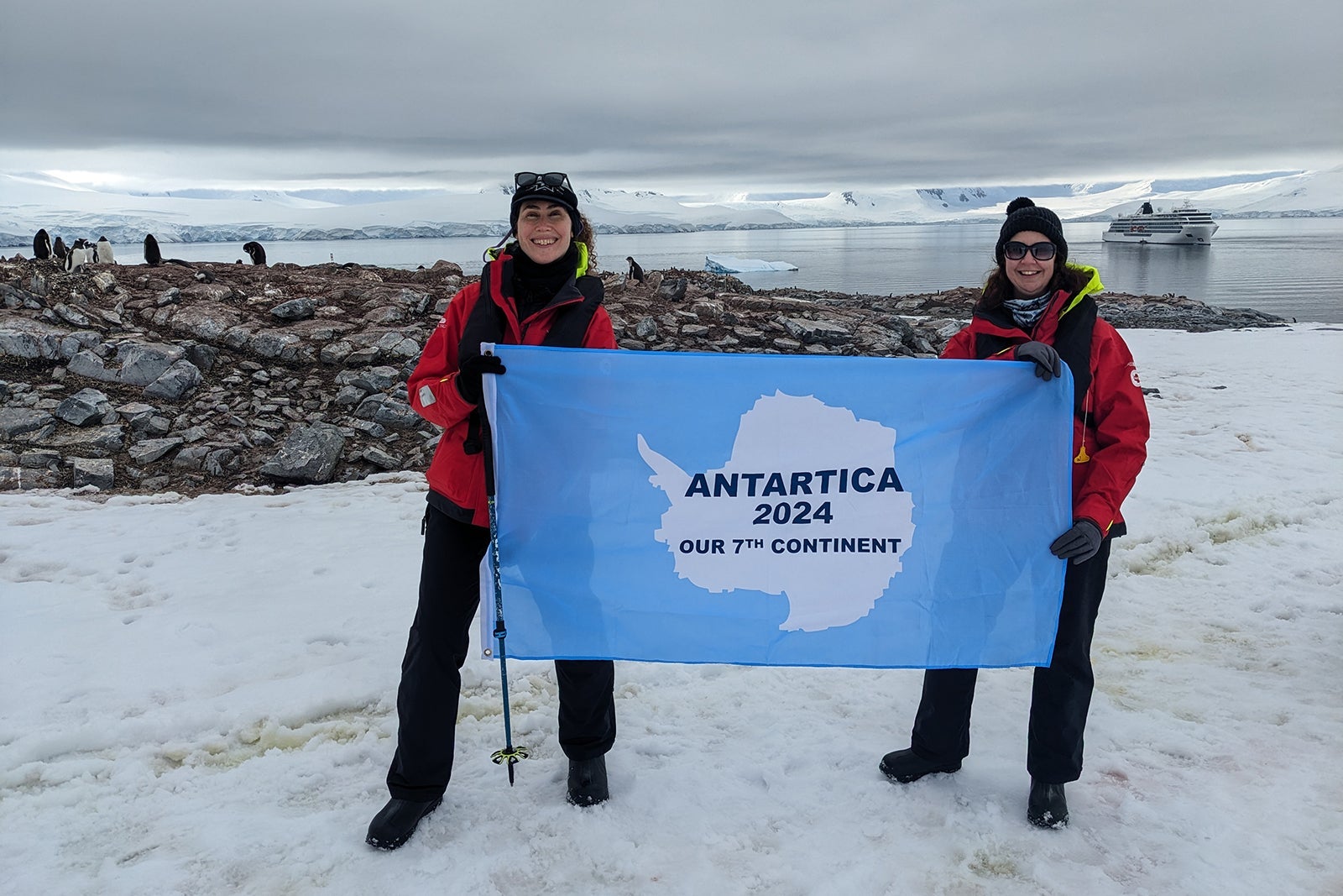 Two women with blue Antarctica flag, posing with penguins behind them