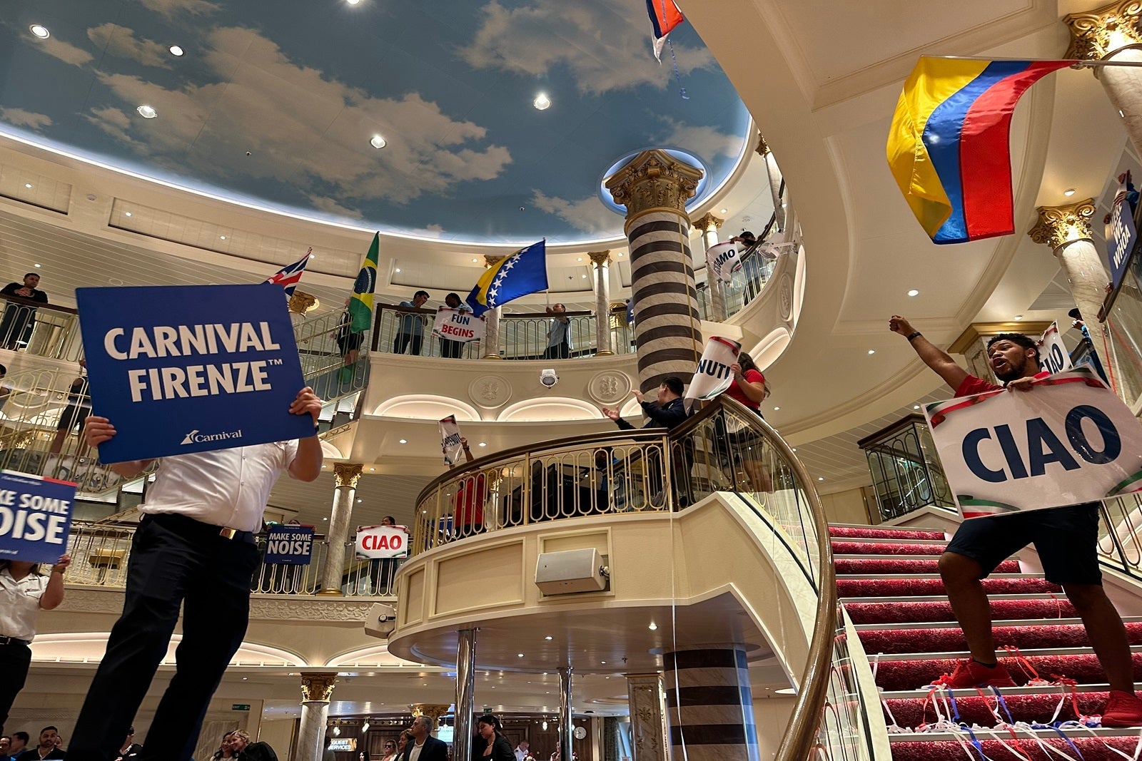 Crew members on a cruise ship wave flags and signs in the atrium to welcome passengers aboard the Carnival Firenze