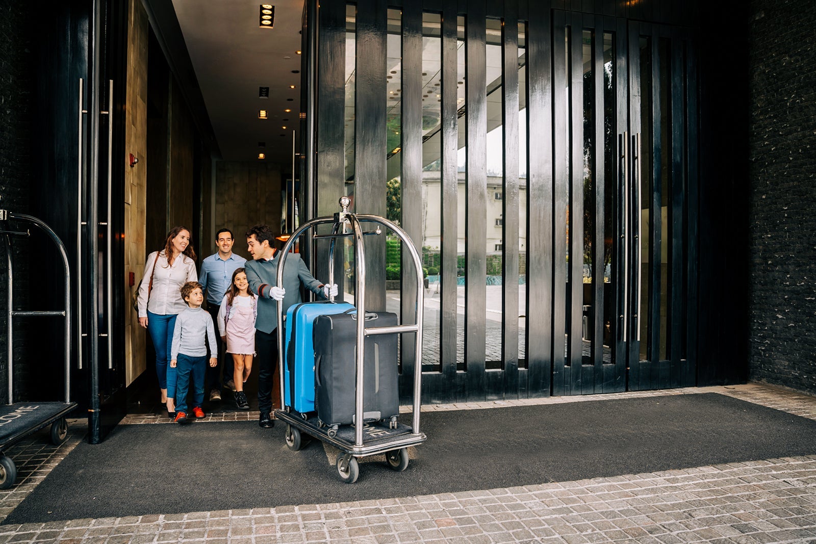 Couple with their two children walking out of luxury hotel following bellhop pushing a luggage cart ready to leave