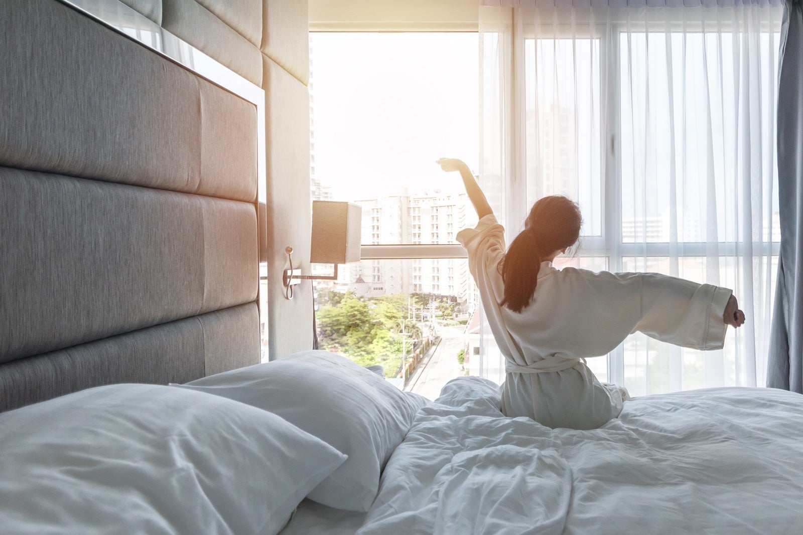 A woman in a hotel room wearing a robe and stretching in front of a window after waking up