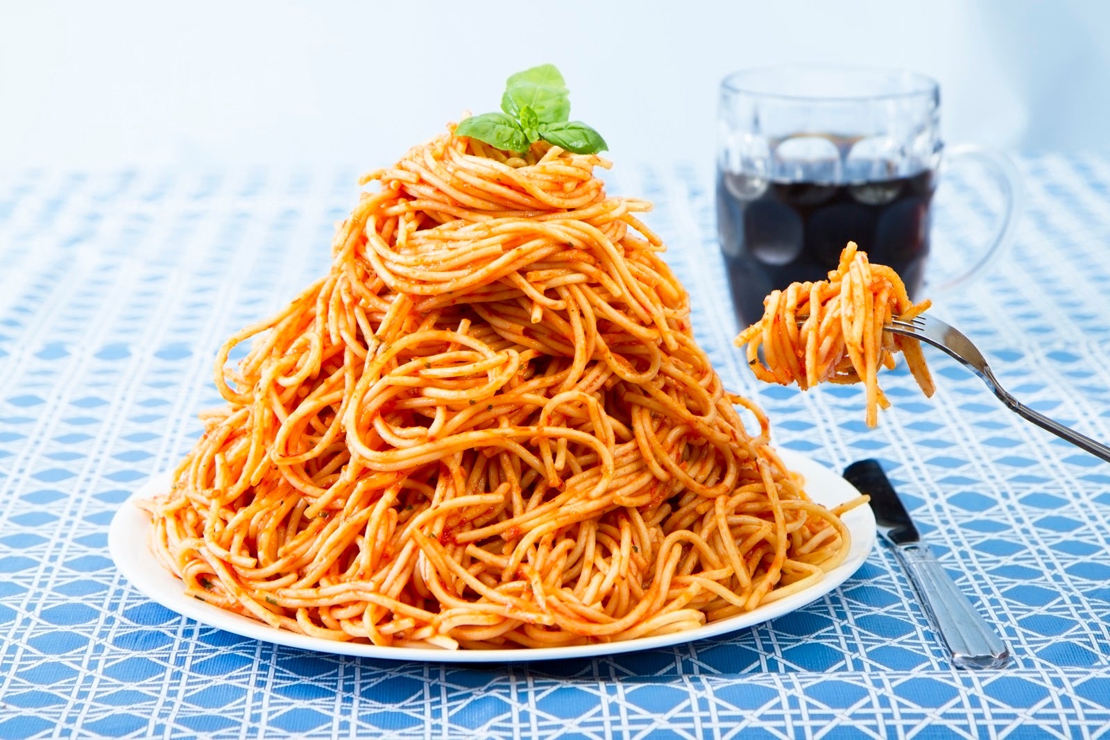 A white plate piled high with spaghetti on a blue tablecloth with a drink and a forkful of spaghetti off to the side