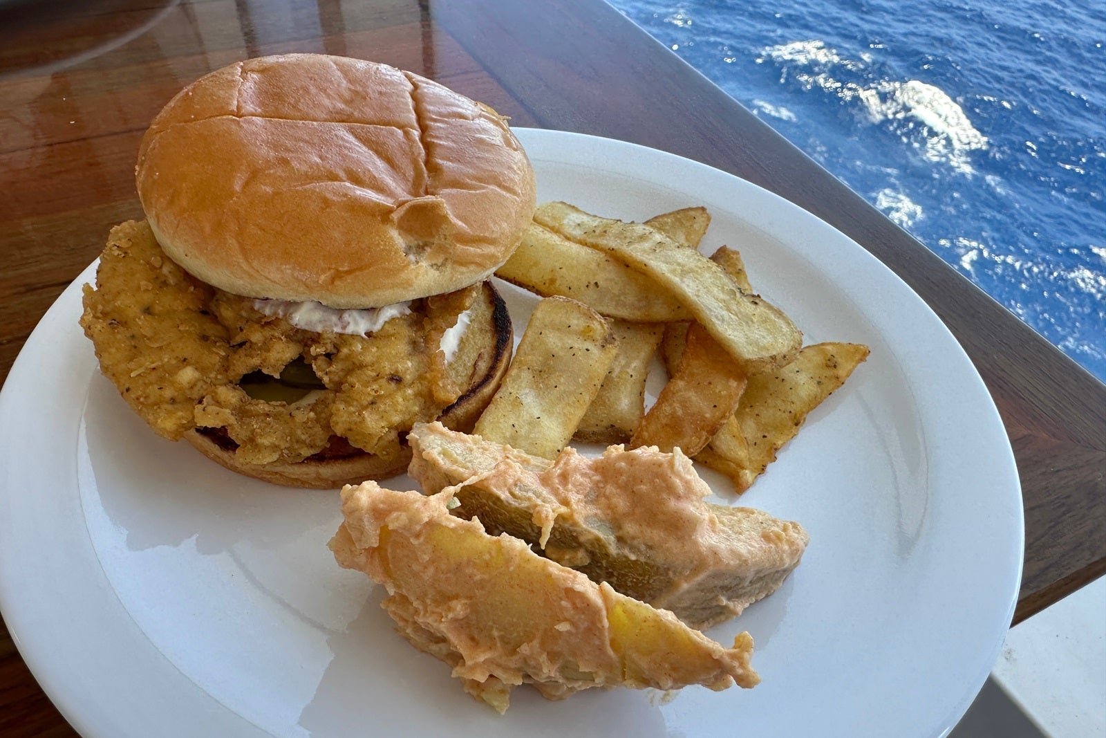 A plate with a chicken sandwich by a window with the ocean below