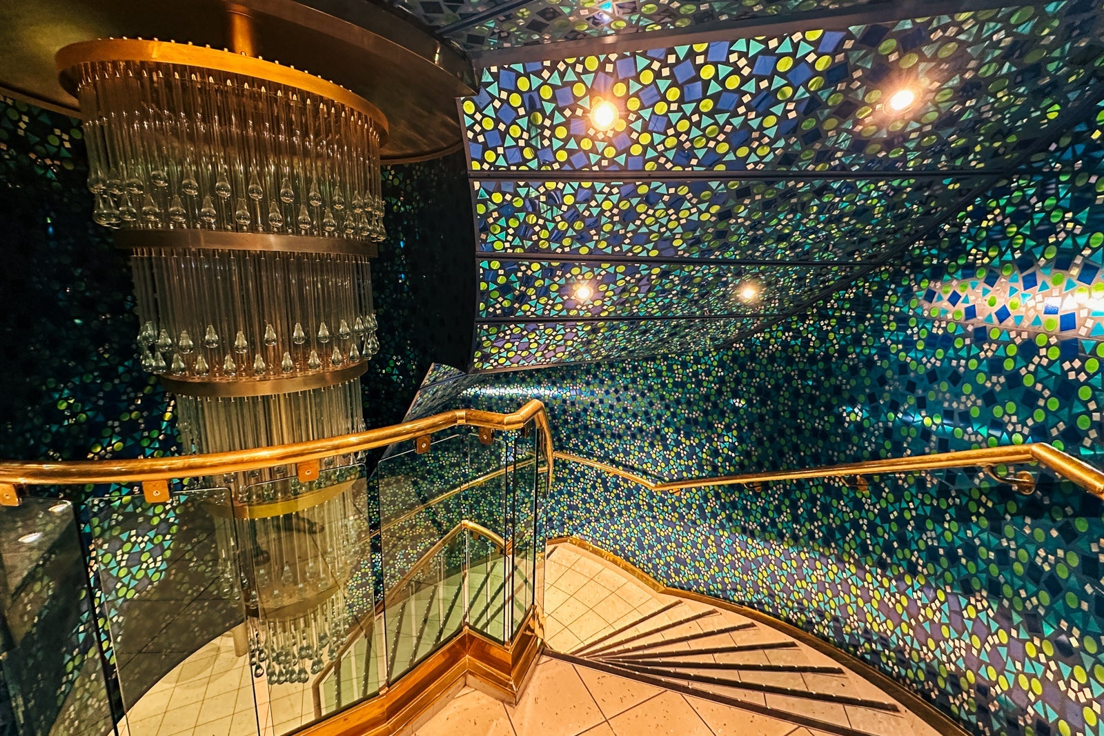 A spiral staircase with a chandelier at the top and blue-green mirrored mosaic walls