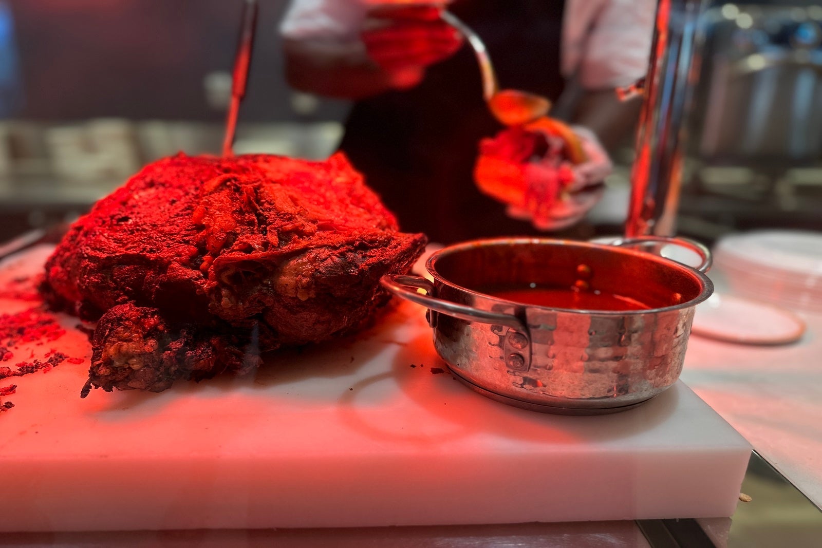 A cut of beef and a stainless steel bowl of jus wait at a carving station while a chef adds jus to a sandwich
