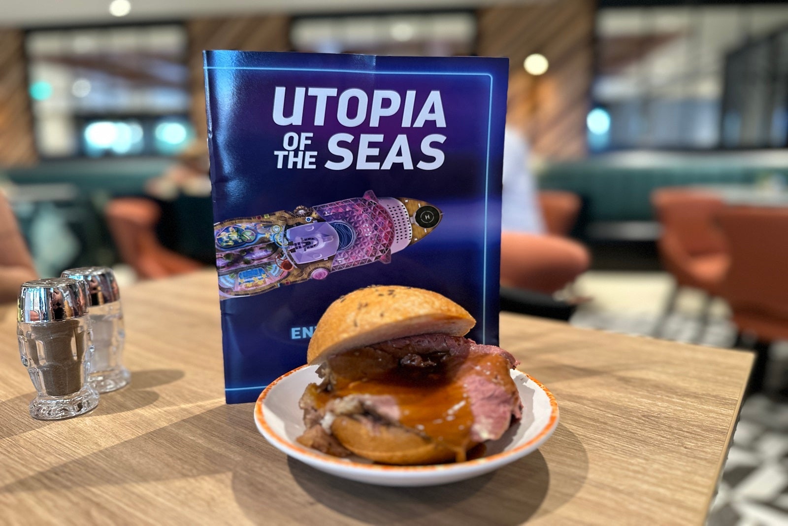 A Royal Kummelweck roast beef sandwich on a plate on a table in front of a Utopia of the Seas brochure