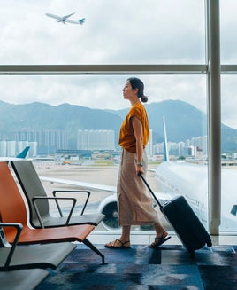 Young Asian woman carrying suitcase, walking by the window at airport terminal. Young Asian female traveller waiting for boarding at airport. Business travel. Travel and vacation concept