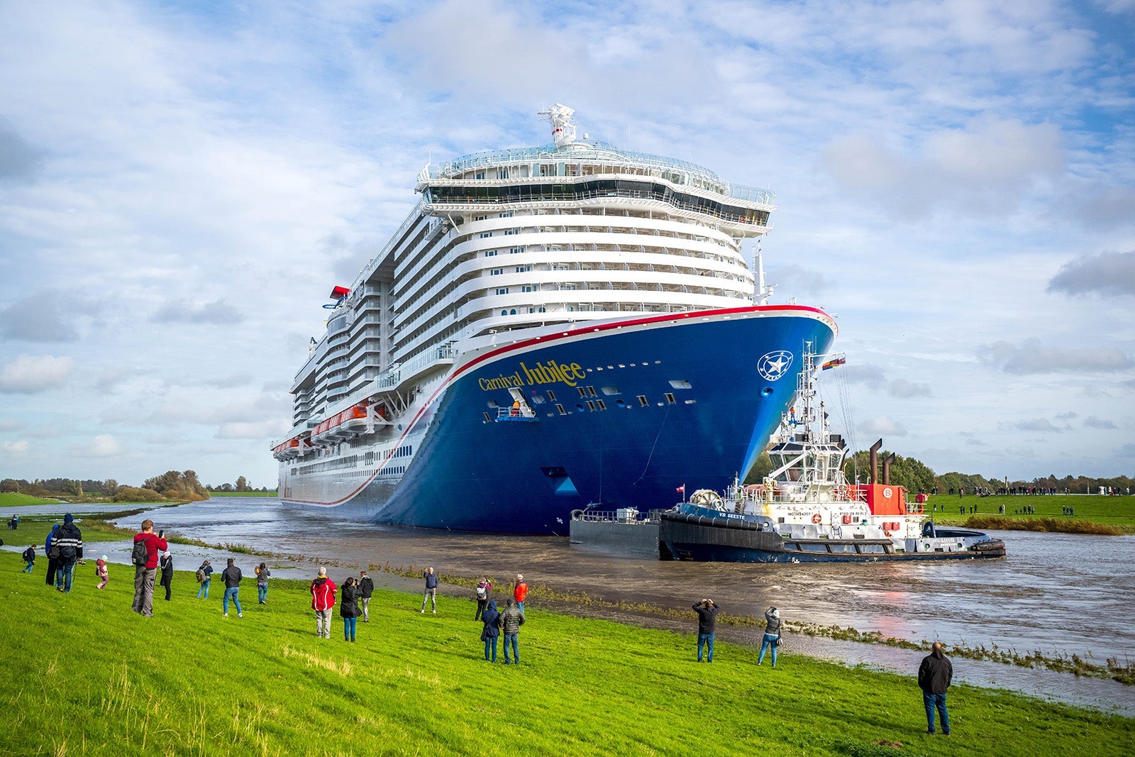 A cruise ship with a blue hull docked near a grassy field where people are watching it