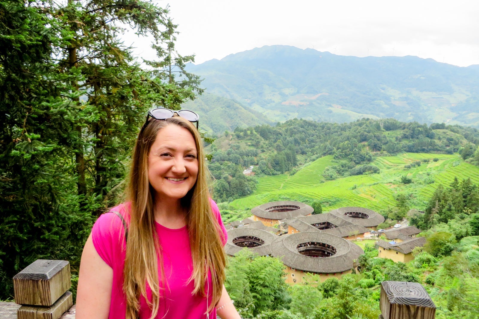 A woman in a pink shirt with sunglasses on her head standing in front of Chinse tulou