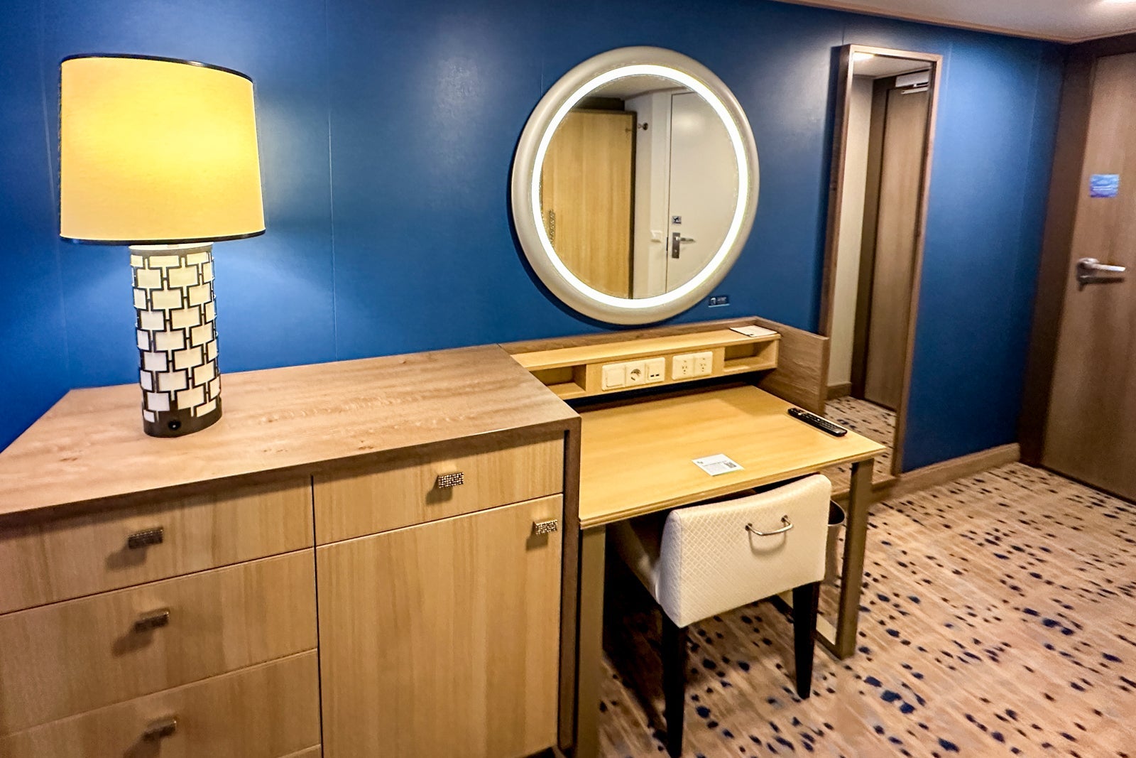 A cruise ship cabin with a vanity, chair, lamp and round mirror with makeup lighting