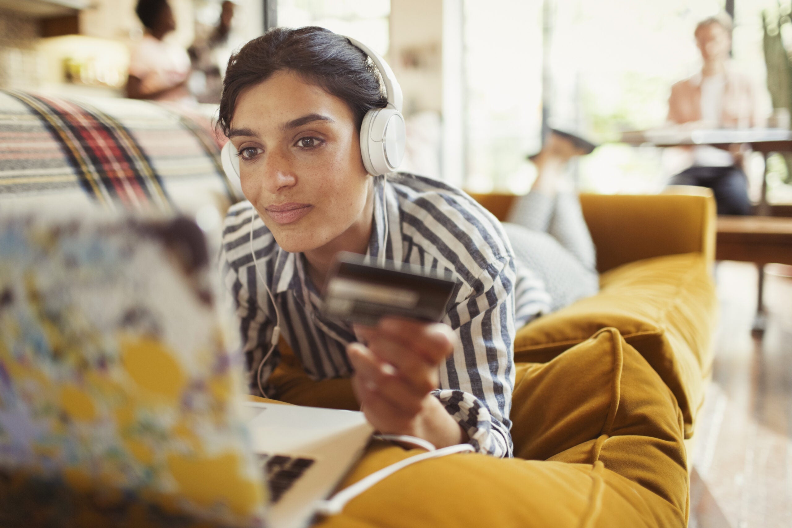 a woman in a striped shirt on a sofa holds a credit card while looking at a laptop
