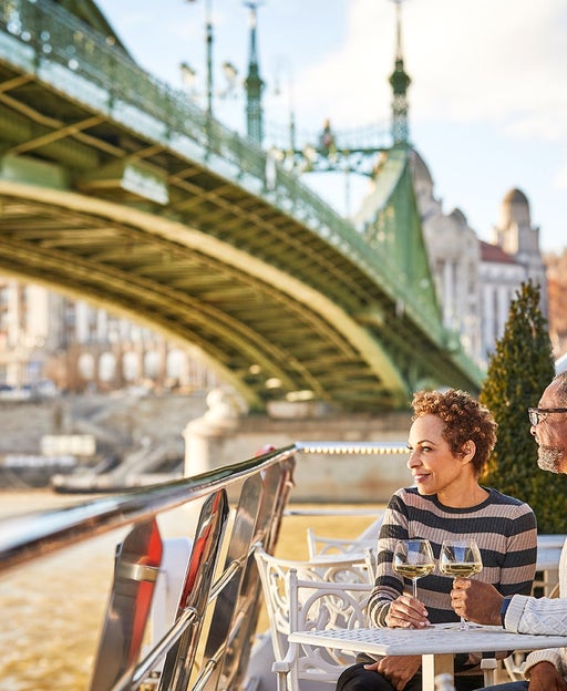 A couple sitting on the deck of a river cruise with drinks as the boat goes under a bridge