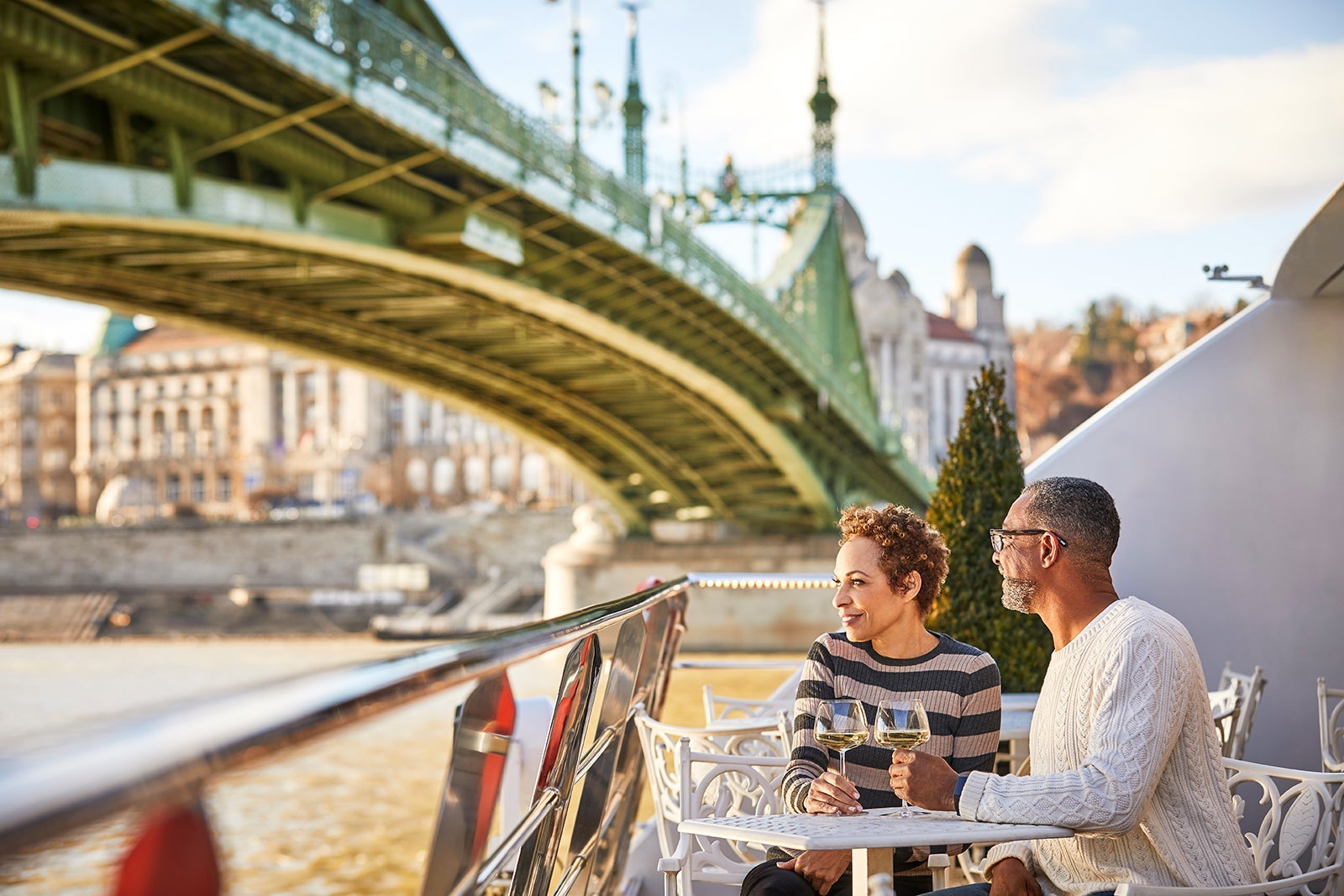 A couple sitting on the deck of a river cruise with drinks as the boat goes under a bridge
