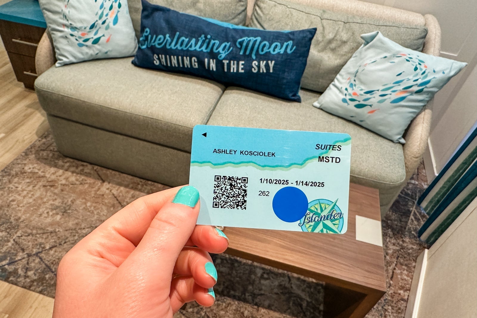 A hand with blue nails holding a cruise keycard in front of a cabin sofa and table