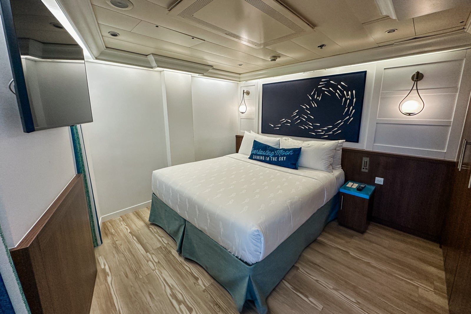 A cruise ship cabin with a king-sized bed, nightstands, lights a wall-mounted TV, pillows and a fish-themed headboard