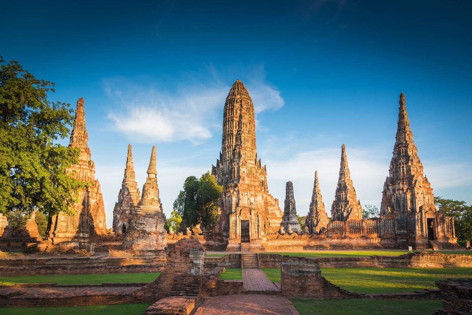 Pointy tan-colored stone temples in Thailand, surrounded by green grass and blue sky