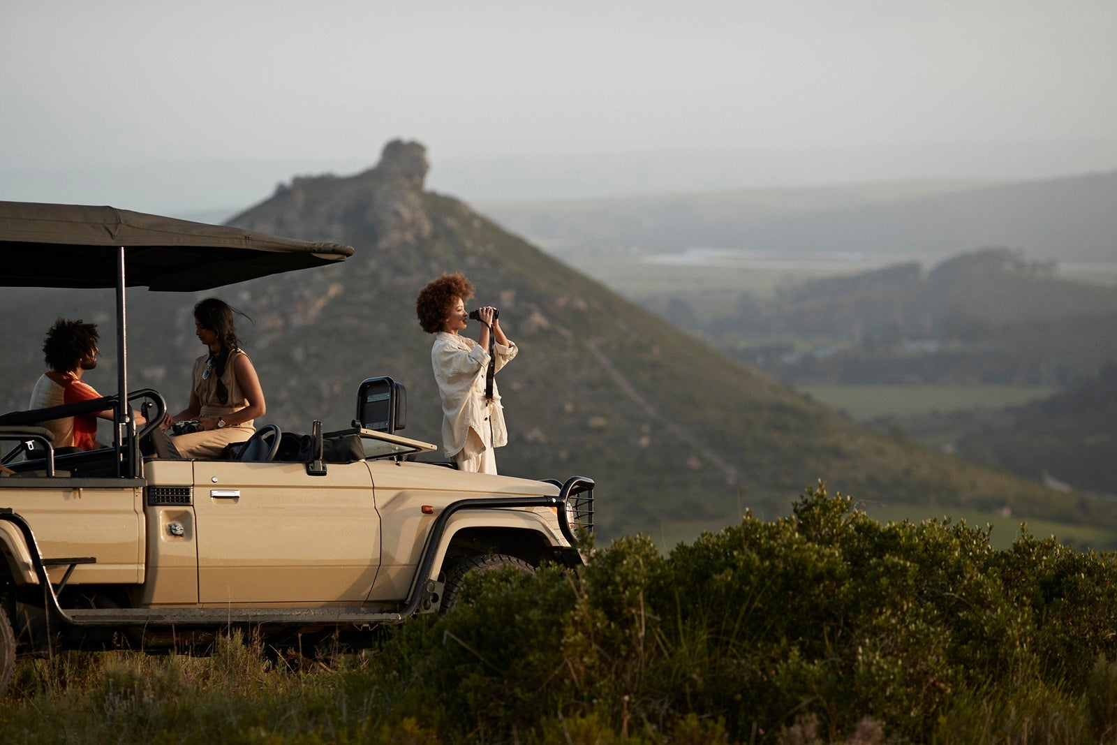 Young woman looking through binoculars while standing near friends in safari vehicle in wildlife park against sky during sunset