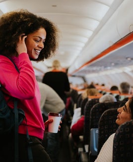A young woman boards a small commercial airplane, carrying a backpack. She walks down the middle aisle, and speaks to a fellow passenger, who is already seated. The two have a friendly exchange.