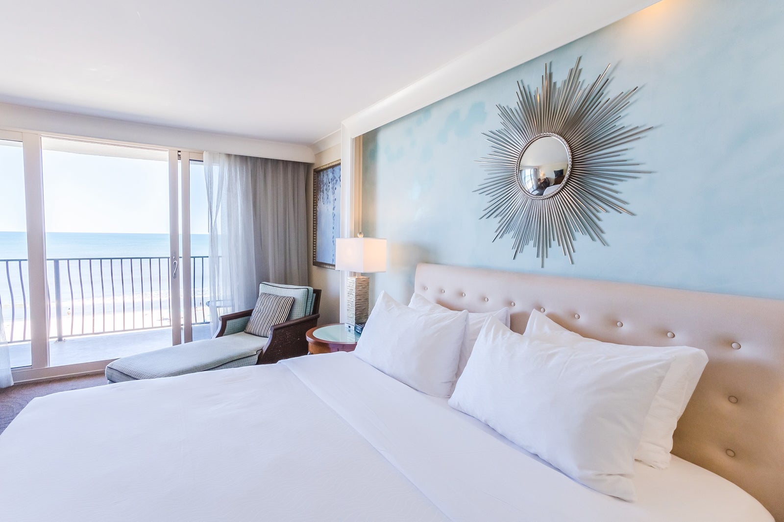 Hotel room with bed all in white sheets and lounge chair with a balcony facing the sea