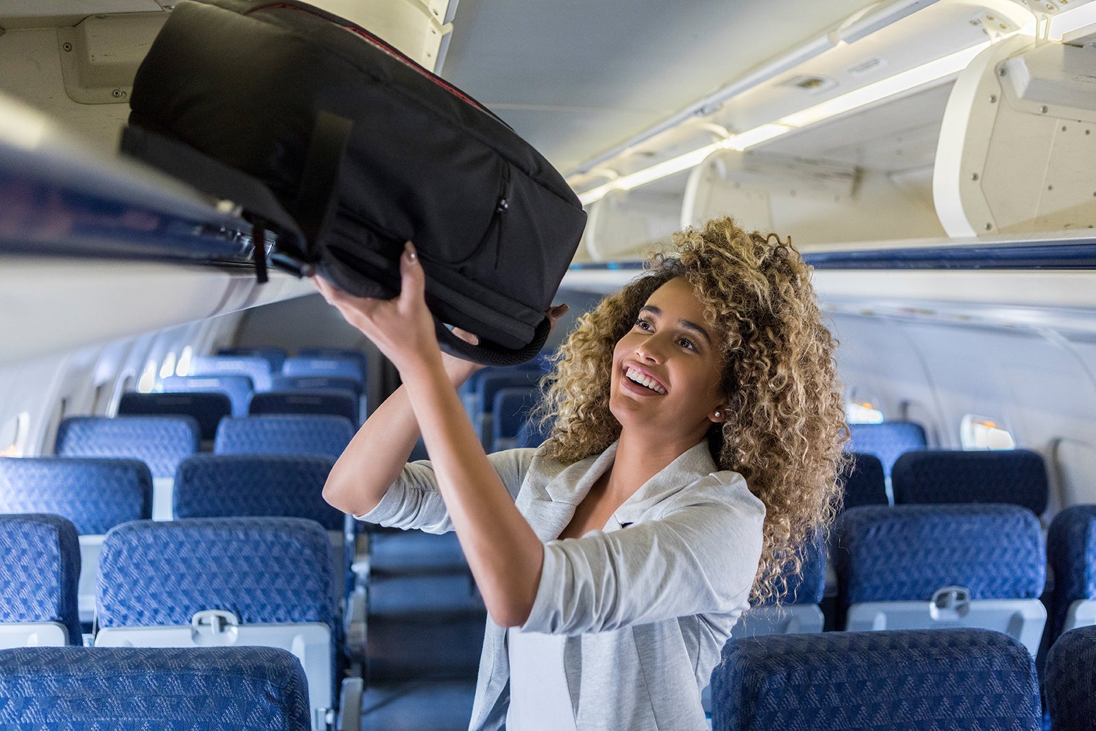 A young woman lifts up her baggage into overhead storage on a plane