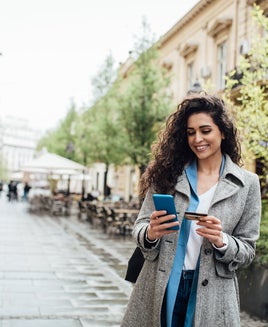 A woman walking around a city with her phone and a credit card in her hands