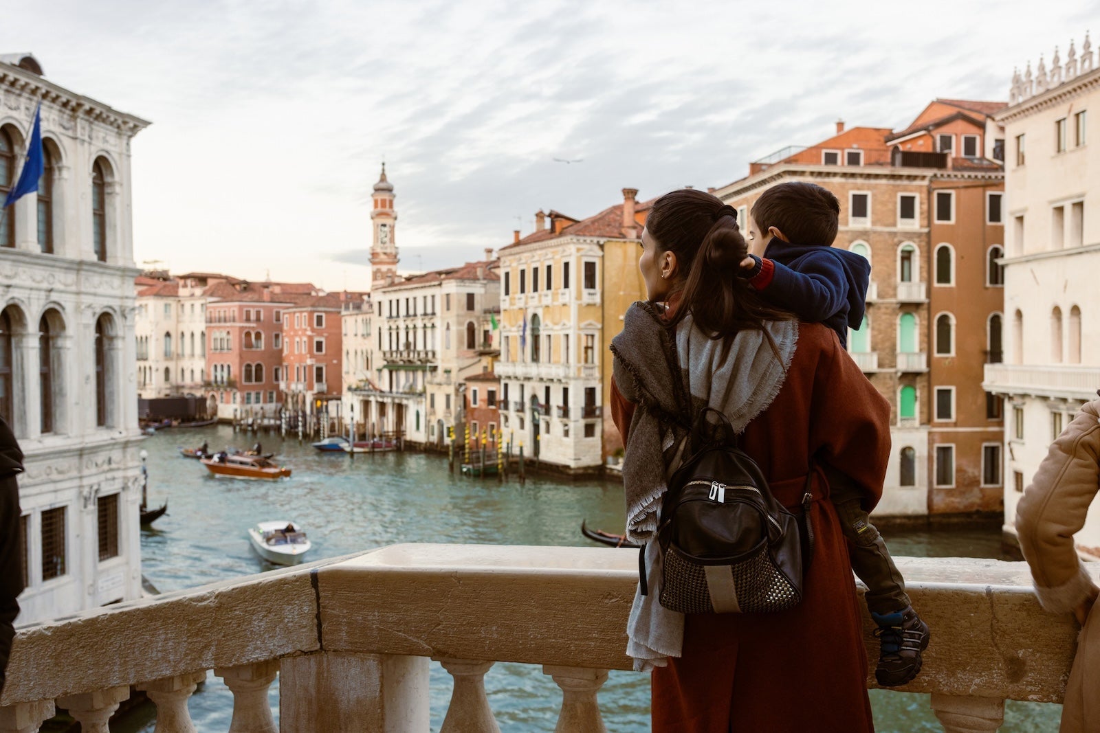 Mother is carrying her son as they enjoy a tourist visit to Venice. They are standing on a bridge overlooking the Grand Canal, with gondolas and boats passing by. The magnificent architecture and stunning waterways of Venice are on full display, providing a picturesque backdrop for their memorable trip. Venice, Italy
