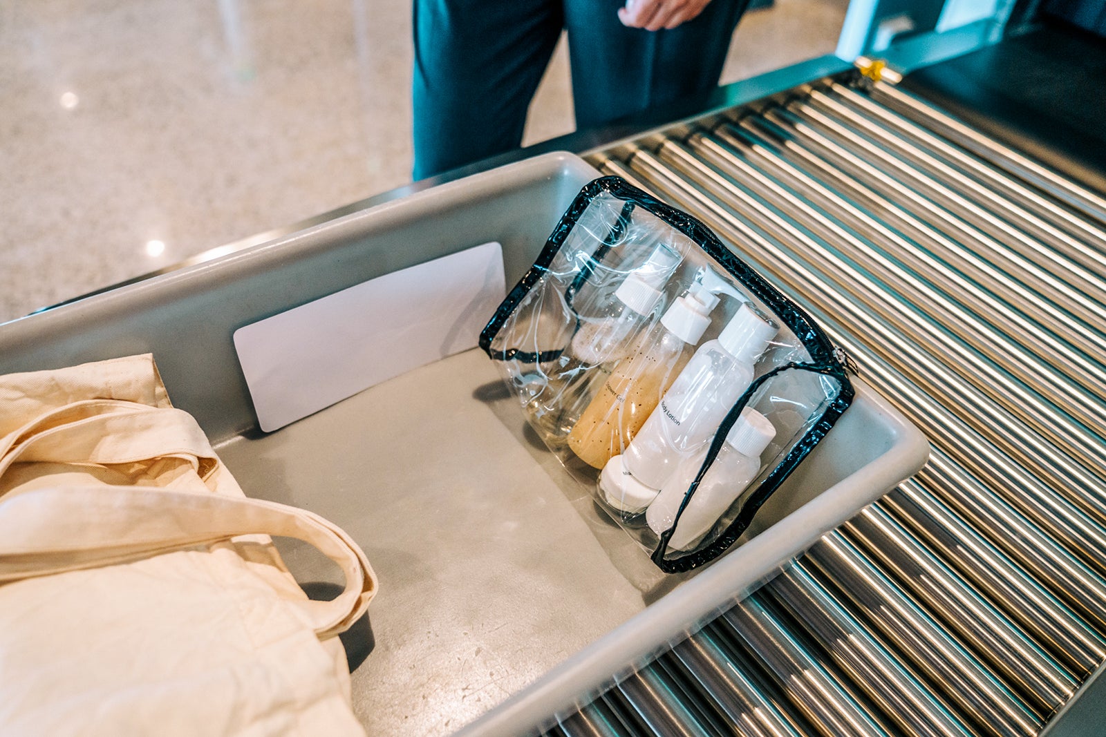 Toiletry bag full of liquids and a tote bag on a plastic tray moving along a roller conveyor at airport security baggage scanner.